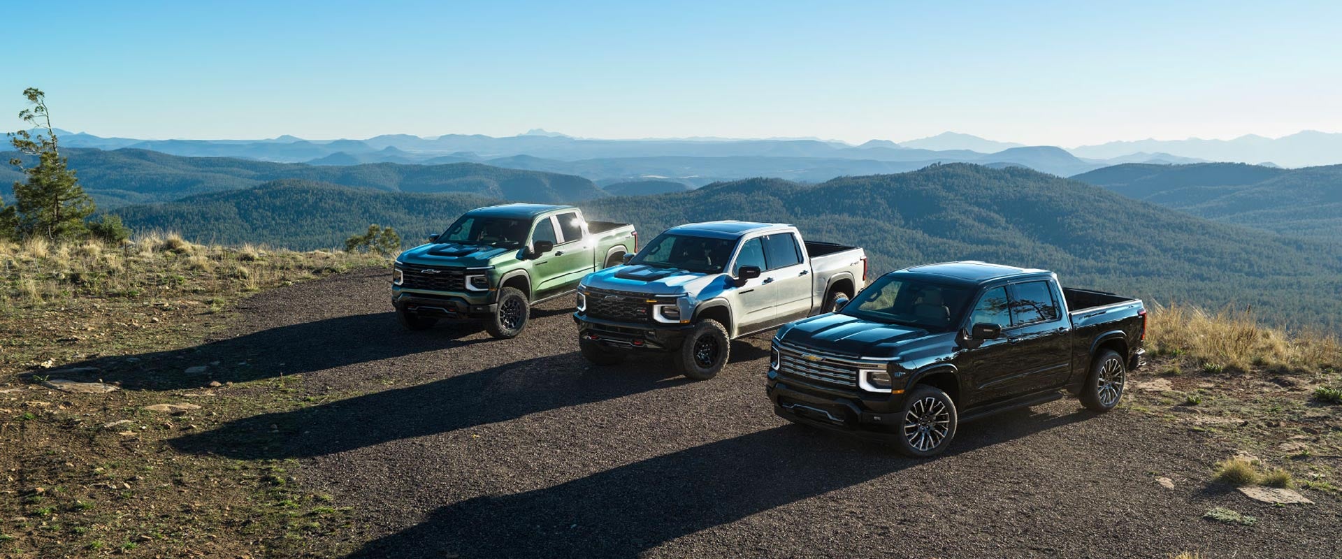 Tres camionetas Chevrolet Silverado 1500 del año 2027 (verde, negra) aparcadas en fila en un campo de tierra, con una cordillera al fondo.